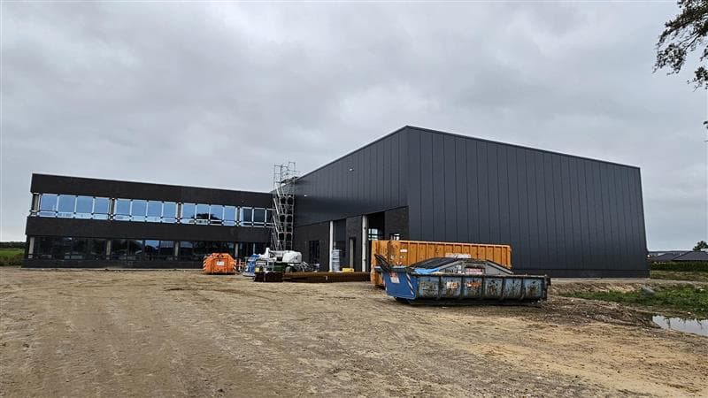 Modern industrial building under construction with dark exterior, scaffolding, and construction materials on a dirt lot under a cloudy sky.